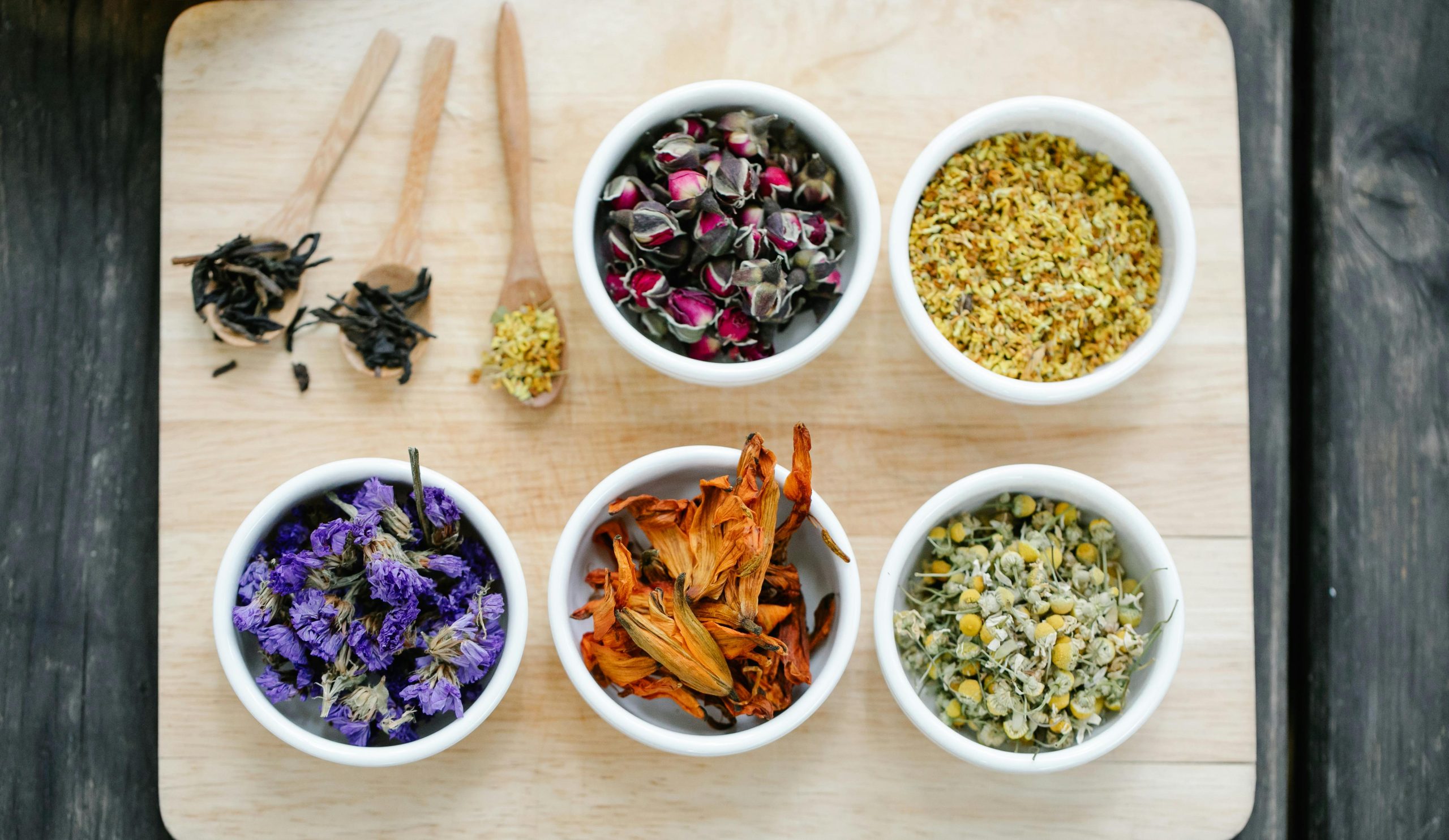 Colorful dried flowers and herbs displayed in bowls on a wooden board.